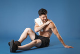 Full length portrait of sick crying fitness guy bare-chested sportsman isolated on blue background studio portrait. Workout sport motivation concept. Sitting on floor get injured put hand on shoulder.