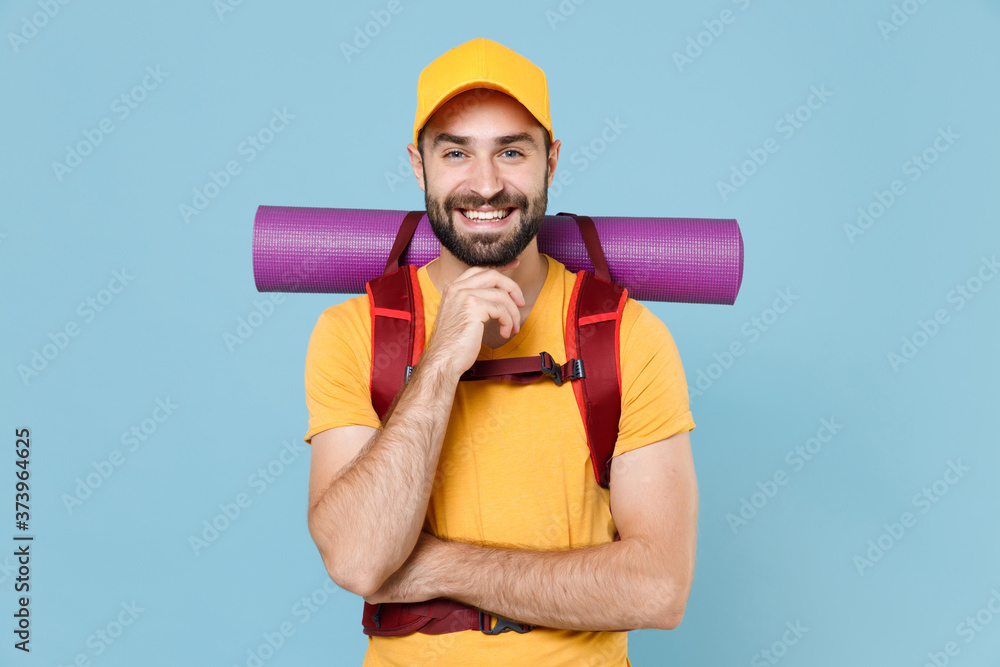 Smiling traveler young man in yellow casual t-shirt, cap with backpack isolated on blue background. Tourist traveling on weekend getaway. Tourism discovering hiking concept. Put hand prop up on chin. Smiling traveler young man in yellow casual t-shirt, cap with backpack isolated on blue background. Tourist traveling on weekend getaway. Tourism discovering hiking concept. Put hand prop up on chin.