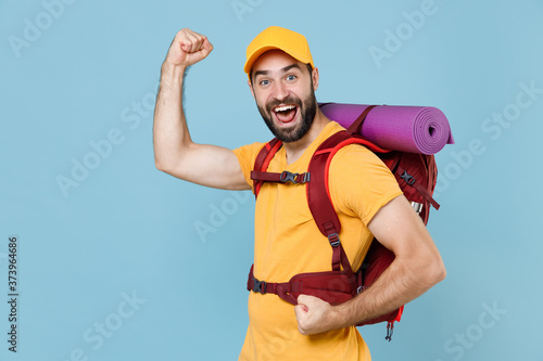 Joyful traveler young man in yellow casual t-shirt cap with backpack isolated on blue background. Tourist traveling on weekend getaway. Tourism discovering hiking concept. Clenching fists like winner.