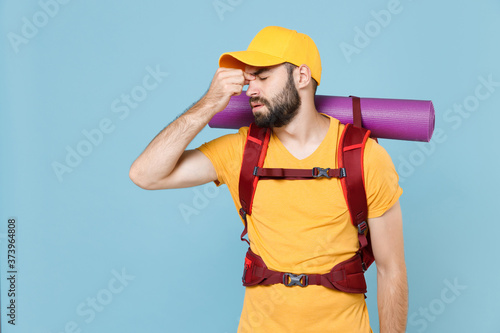 Tired traveler young man in yellow t-shirt cap with backpack isolated on blue background. Tourist traveling on weekend getaway. Tourism discovering hiking concept. Keep eyes closed, put hand on nose.