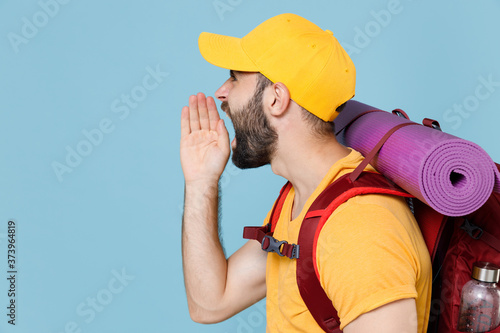 Side view of angry traveler young man in cap backpack isolated on blue background. Tourist traveling on weekend getaway. Tourism discovering hiking concept. Screaming with hand gesture near mouth.