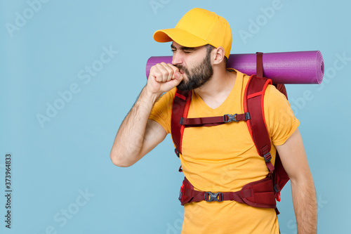 Sick traveler young man in yellow t-shirt cap backpack isolated on blue background. Tourist traveling on weekend getaway. Tourism discovering hiking concept Coughing sneezing covering mouth with hand.