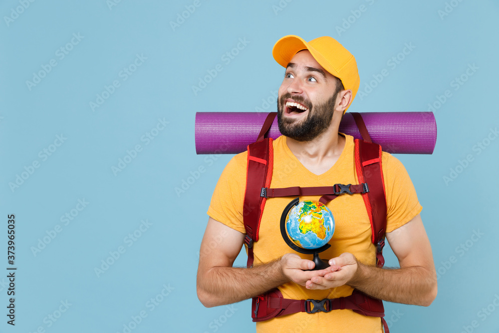 Excited young traveler man in yellow t-shirt cap with backpack isolated on blue background. Tourist traveling on weekend getaway. Tourism discovering hiking concept. Hold world globe looking aside up.