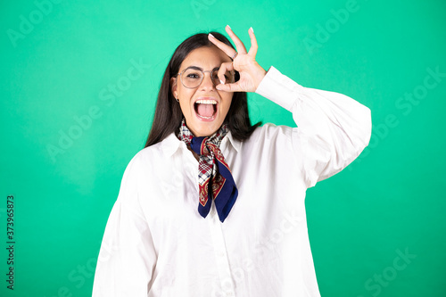 Young beautiful business woman over isolated green background doing ok gesture shocked with smiling face, eye looking through fingers