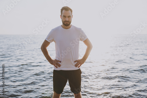 Portrait of young handsome bearded athletic man guy 20s in casual white t-shirt posing training warming up standing with arms akimbo on waist looking camera at sunrise over the sea outdoors.