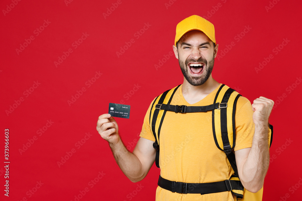 Delivery man in yellow cap t-shirt uniform thermal bag backpack with food hold credit card isolated on red background studio Guy male employee work courier Service coronavirus covid-19 virus concept