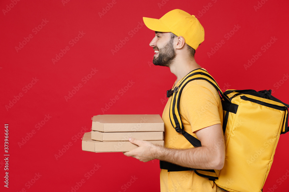 Side view delivery man in yellow cap t-shirt thermal bag backpack with food hold pizza in cardboard flatbox isolated on red background studio Employee work courier Service coronavirus covid-19 concept Side view delivery man in yellow cap t-shirt thermal bag backpack with food hold pizza in cardboard flatbox isolated on red background studio Employee work courier Service coronavirus covid-19 concept