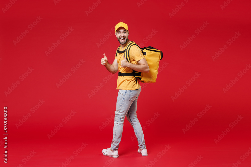 Full length back view delivery employee man guy male 20s in yellow cap t-shirt uniform thermal food bag backpack work courier service in quarantine covid-19, standing isolated on red background studio Full length back view delivery employee man guy male 20s in yellow cap t-shirt uniform thermal food bag backpack work courier service in quarantine covid-19, standing isolated on red background studio