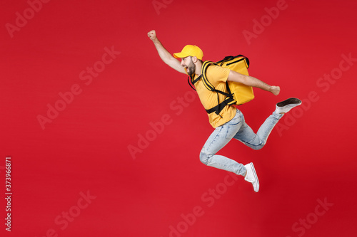 Fototapeta Naklejka Na Ścianę i Meble -  Full length delivery employee man guy male 20s in yellow cap t-shirt uniform thermal food bag backpack work courier service during quarantine covid-19 virus jumping isolated on red background studio.
