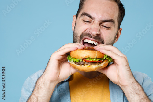 Close up of crazy young bearded man guy 20s wearing casual clothes posing holding eating american classic fast food burger looking aside isolated on pastel blue color wall background studio portrait.