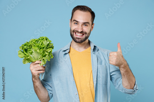 Smiling young bearded man guy 20s wearing casual clothes posing holding bunch of fresh greens salad showing thumb up looking camera isolated on pastel blue color wall background studio portrait.