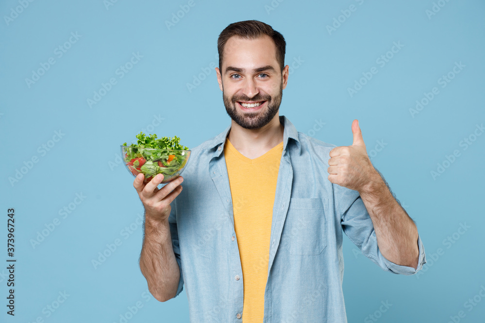 Smiling young bearded man guy 20s wearing casual clothes posing holding in hand vegetable salad in glass bowl showing thumb up looking camera isolated on pastel blue color background studio portrait. Smiling young bearded man guy 20s wearing casual clothes posing holding in hand vegetable salad in glass bowl showing thumb up looking camera isolated on pastel blue color background studio portrait.