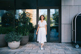 Positive woman in stylish dress walking away from doorway of modern building