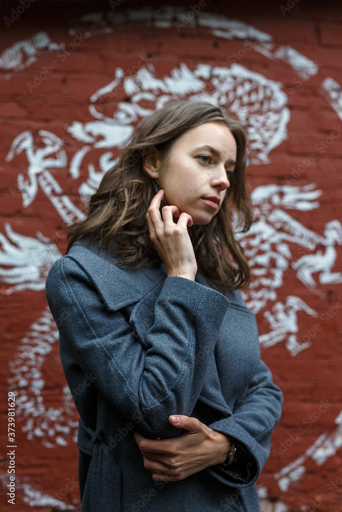 Fototapeta premium young thoughtful pretty girl in gray coat and turtleneck poses in front of red brick wall with graffiti. Street style portrait of female, urban photoshoot of elegant model