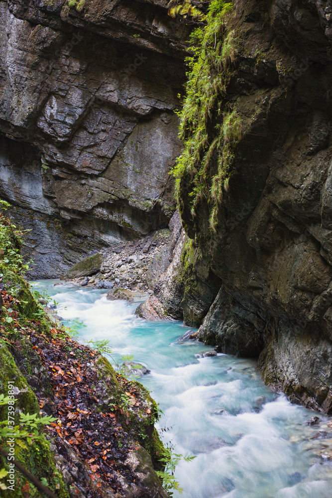 A wild mountain river with clear blue water flows through the open area of the Partnachklamm gorge in Germany. The rapid stream slides over the rocks.
