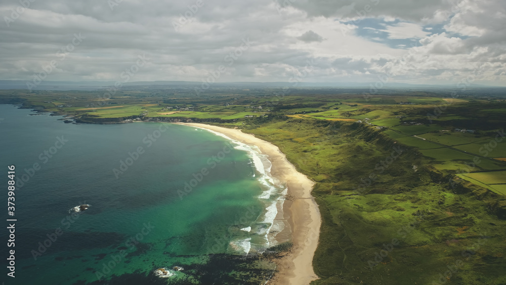 Aerial view sandy coastline, green grass meadows. Waves crashing out of shore and come back to ocean. Atlantic bay serene scenery in dusk summer daytime cinematic shot