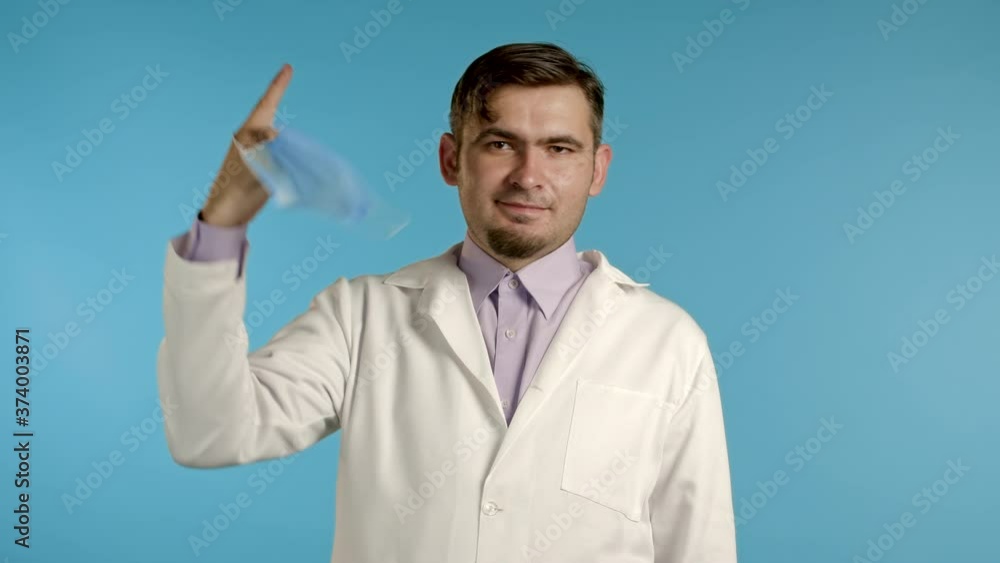 Pleased man in medical coat twists removed protective mask on finger and throws it on floor as symbol of pandemic, epidemic end. Blue studio background. Pleased man in medical coat twists removed protective mask on finger and throws it on floor as symbol of pandemic, epidemic end. Blue studio background.