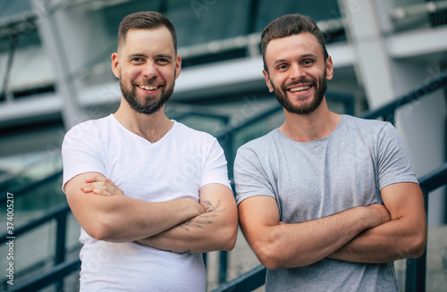 Two happy guys with crossed arms. Two young bearded handsome friends in t-shirts are posing together on the city background.