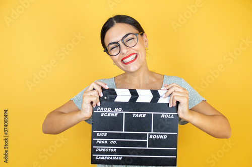 Young beautiful woman wearing glasses standing over isolated yellow background holding clapperboard very happy