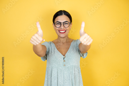 Young beautiful woman wearing glasses standing over isolated yellow doing happy thumbs up gesture with hands. Approving expression looking at the camera showing happiness background photo
