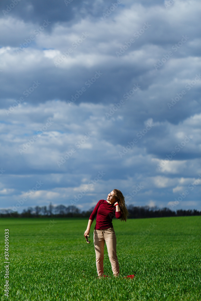 Beautiful young blonde woman stands on green grass in a park. Blue sky with clouds. The girl smiles and enjoys a good day. Beautiful young blonde woman stands on green grass in a park. Blue sky with clouds. The girl smiles and enjoys a good day.