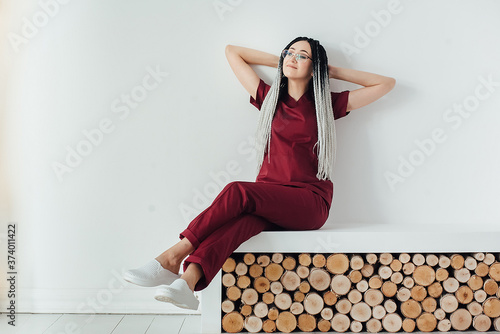 a young woman with African braids on her head, in a red medical uniform, sits with her hands behind her head and is resting. she has a break
