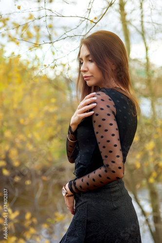 Portrait of pretty young woman of Slavic appearance in dark dress in autumn, standing against background of an autumn Park and pond with water. Cute model walks in Park in golden autumn