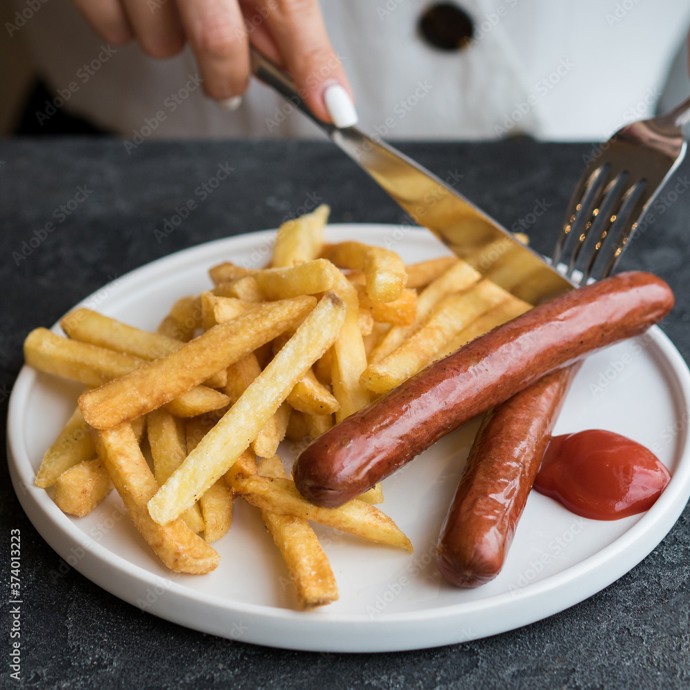 Two sausages and french fries with ketchup on a white plate. The girl cuts the food with a knife and fork. Only hands are visible. Lunch at a cafe. Junk food. Fast food. Dinner at the cafe. Two sausages and french fries with ketchup on a white plate. The girl cuts the food with a knife and fork. Only hands are visible. Lunch at a cafe. Junk food. Fast food. Dinner at the cafe.