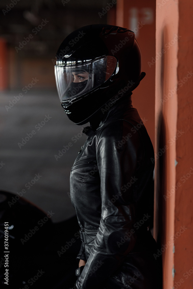Stylish young woman motorcycle rider in black protective gear and full-face helmet near her bike on underground parking. Stylish young woman motorcycle rider in black protective gear and full-face helmet near her bike on underground parking.