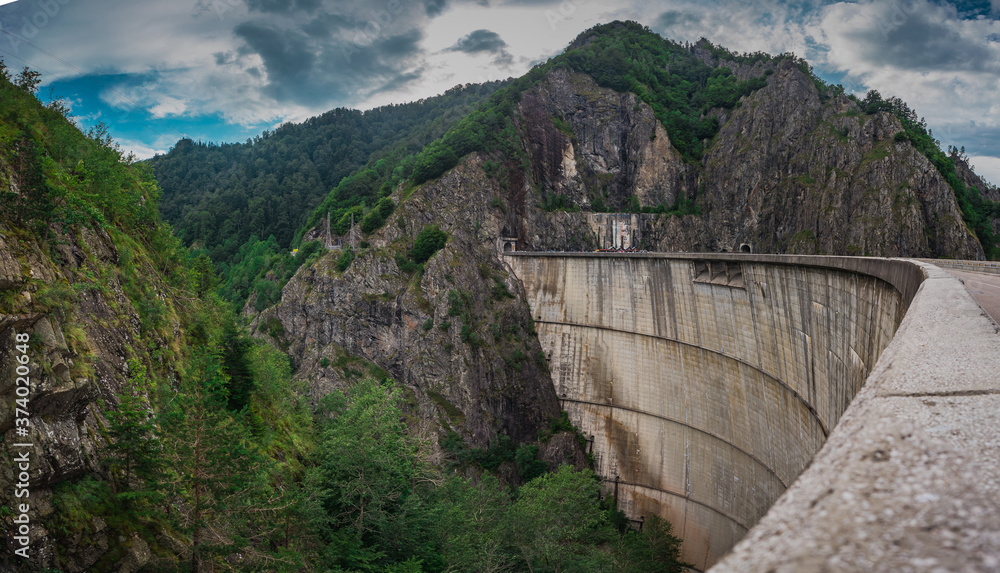 Panoramic photo of Vidraru dam or Barajul Vidraru in Romania, a concrete hydroelectric power plant dam built  in the carpathian mountains on a cloud dull summer day.