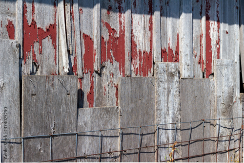 Sunny exterior weathered wood siding on a rustic old 19th Century barn, with peeling red paint and rusty metal fencing