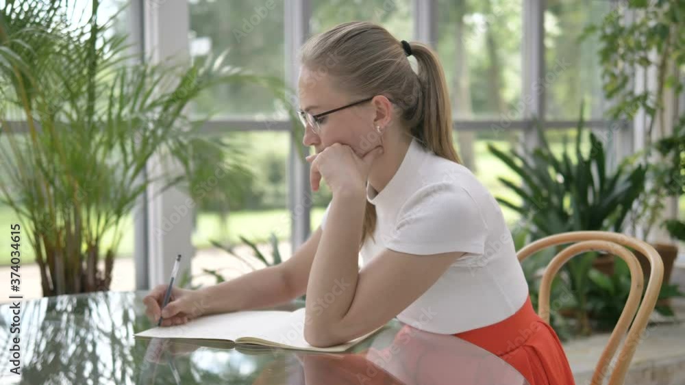 elegant blonde businesswoman in white t-shirt writes in paper notebook with pen sitting at table in cafe with tropical pot plants elegant blonde businesswoman in white t-shirt writes in paper notebook with pen sitting at table in cafe with tropical pot plants