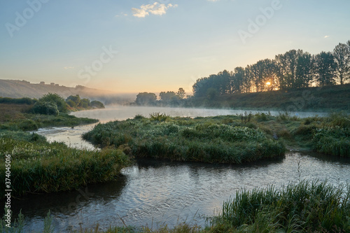 Summer time. Morning dawn over the river in a hazy, thoughtful haze. Beautiful view of the forest and river covered with fog early in the morning.