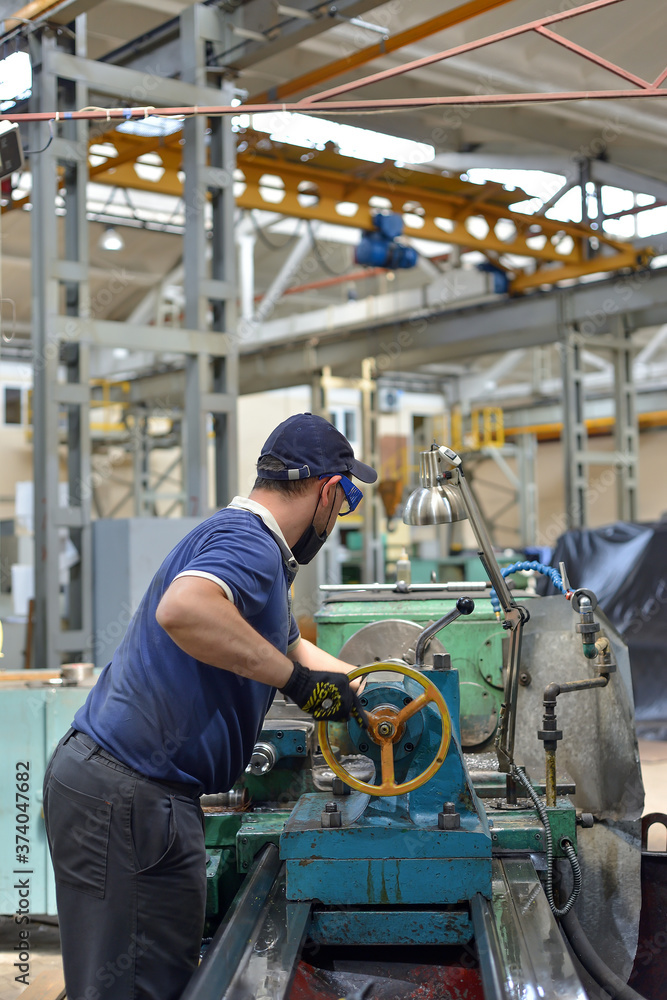 Working turner wearing a protective antiviral mask on a lathe in a workshop. The machine operator at the workplace controls the equipment. Working turner wearing a protective antiviral mask on a lathe in a workshop. The machine operator at the workplace controls the equipment.