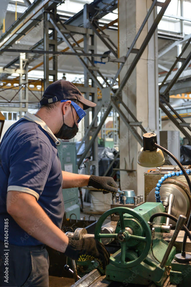 A working turner wearing a protective antiviral mask works on a lathe under quarantine conditions. A working turner wearing a protective antiviral mask works on a lathe under quarantine conditions.