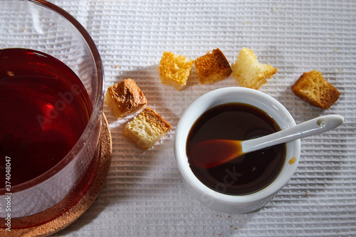 Square toasted pieces of homemade delicious rusk, hardtack, Dryasdust, zwieback, Liquid honey in a saucer and black tee in a cap on a white tablecloth. photo