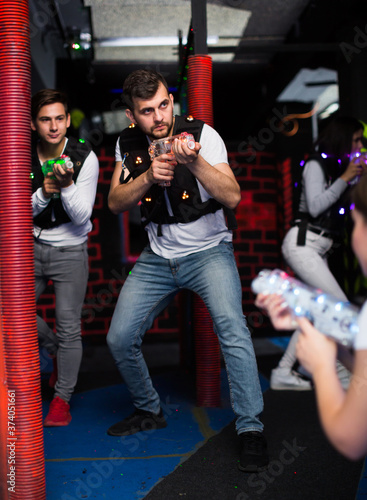 Excited guy aiming laser gun at other players during lasertag game in dark room.