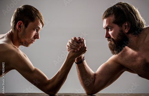 Arms wrestling thin hand, big strong arm in studio. Two man's hands clasped arm wrestling, strong and weak, unequal match. Heavily muscled bearded man arm wrestling a puny weak man photo