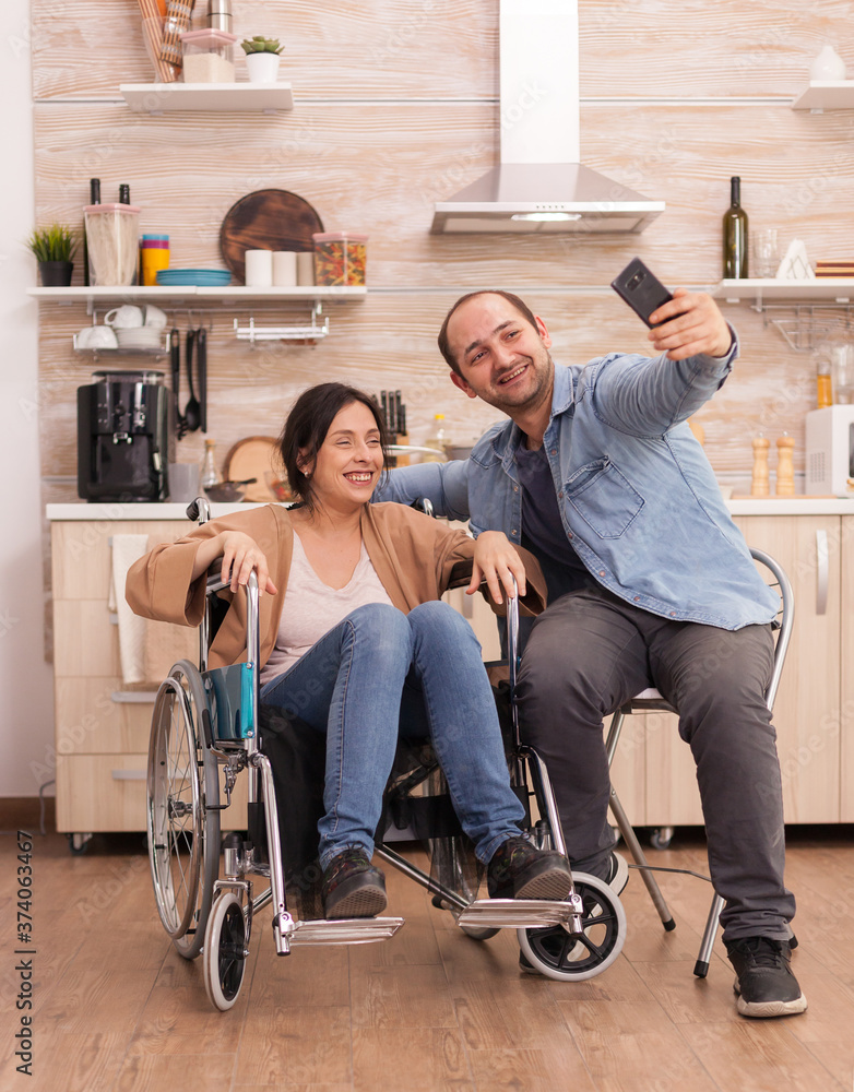 Obraz premium Positive woman in wheelchair and husband taking a selfie in kitchen using smartphone. Disabled paralyzed handicapped woman with walking disability integrating after an accident.