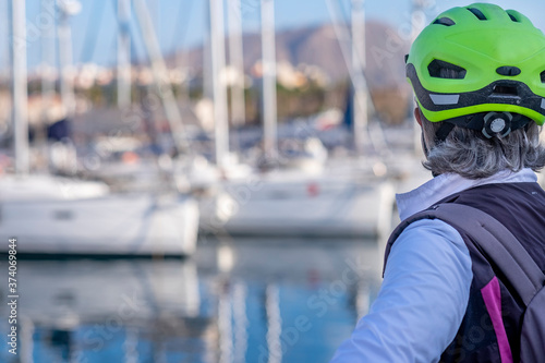 Back view of senior woman with helmet enjoying excursion at sea with her bicycle. Sailboats in the background.