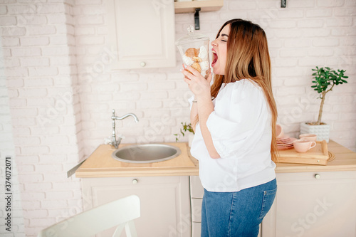 plus size model in jeans and a white t-shirt  holding a sweet  opens her mouth in the kitchen. The body is positive. One beauty concept. XXXL fashion