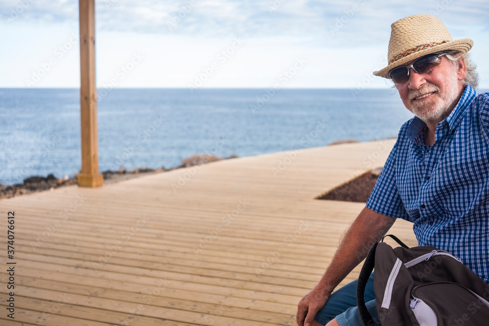 Smiling senior man bearded, white hair and straw hat relaxing on a bench enjoying sea excursion - horizon over water - wooden walkway