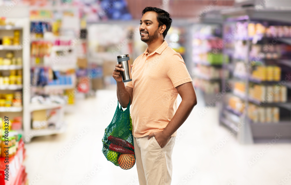 sustainability, consumerism and eco friendly concept - happy indian man holding green reusable string bag for food shopping and tumbler or thermo cup over supermarket or grocery store on background