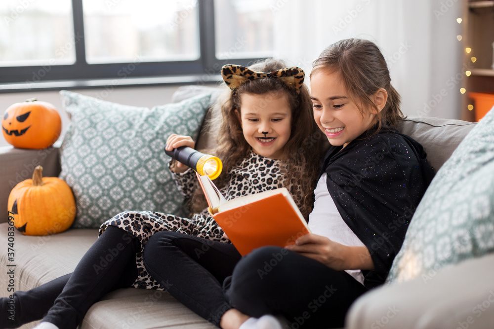 halloween, holiday and childhood concept - little girls in party costumes with flashlight reading book at home halloween, holiday and childhood concept - little girls in party costumes with flashlight reading book at home