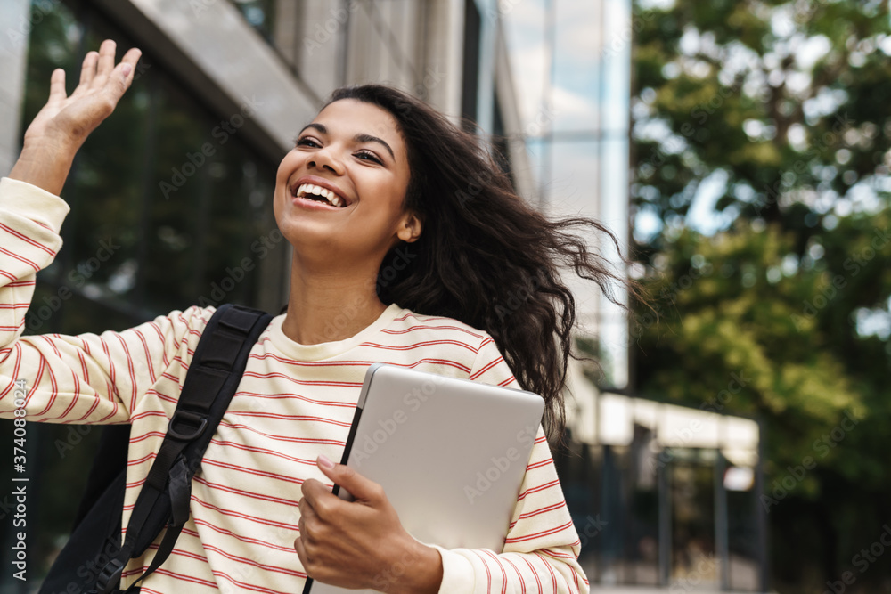 Image of african girl waving hand and holding laptop while walking Image of african girl waving hand and holding laptop while walking