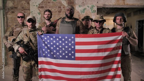 Serious multinational males soldiers in camouflage, bulletproof vests posing with US flag. Brave patriots of USA armed forces standing with weapons and holding symbol of United States of America
