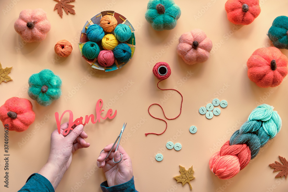 Hands with scissors cutting out word Danke from paper. It means Thank you in German language. Hobby craft materials in Fall colors. Autumn background with felt pumpkins, wool bundle, buttons. Hands with scissors cutting out word Danke from paper. It means Thank you in German language. Hobby craft materials in Fall colors. Autumn background with felt pumpkins, wool bundle, buttons.