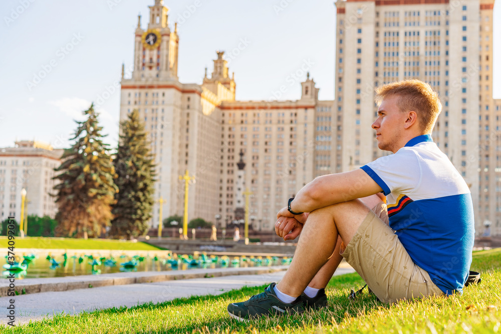 A young blond man sits on the grass in front of the main University of Russia, Moscow high-rise