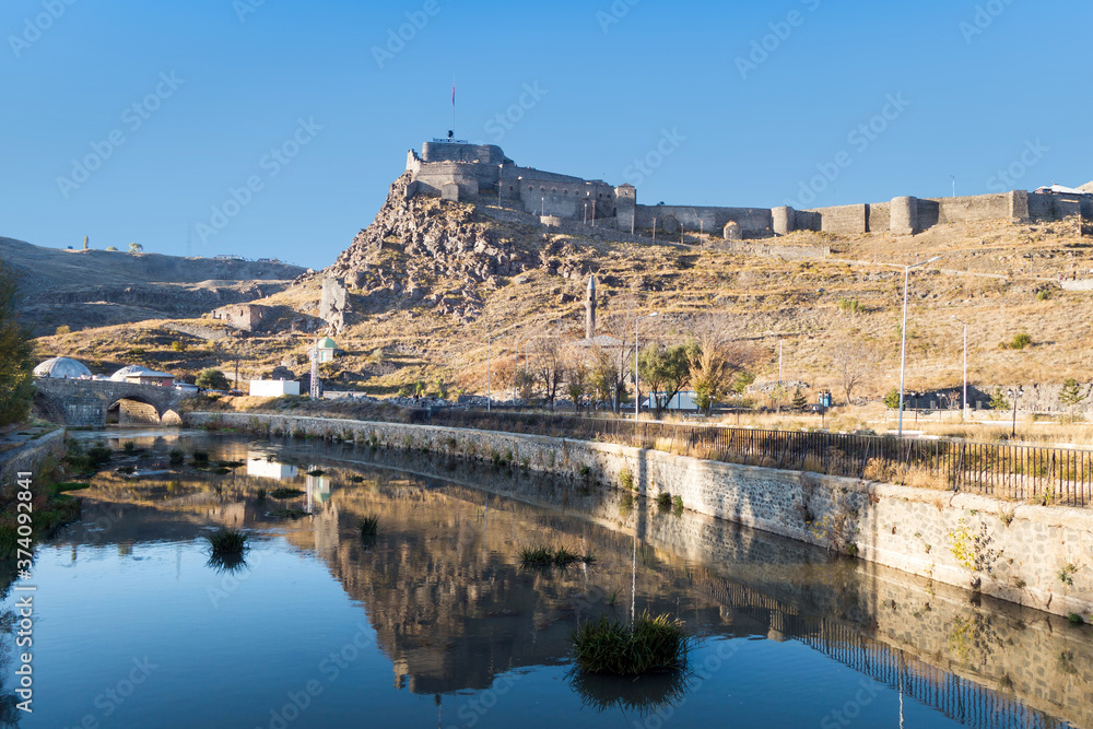 Panoramic view in evening onto tourist attractions of Kars, Turkey: Castle & ancient stone bridge across Kars river. Near flag are portrait of Ataturk & writing in Turkish 'Motherland remembers you'