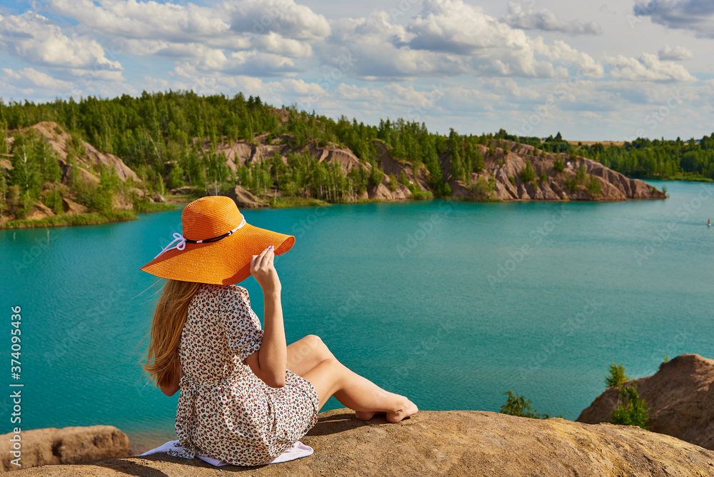 A girl in a hat sits on a high hill and looks at a blue lake.